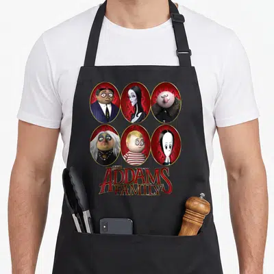 Circle family aprons - Six oval portraits of The Addams Family characters with red and yellow title text below — shown on a black cotton canvas apron against a white background