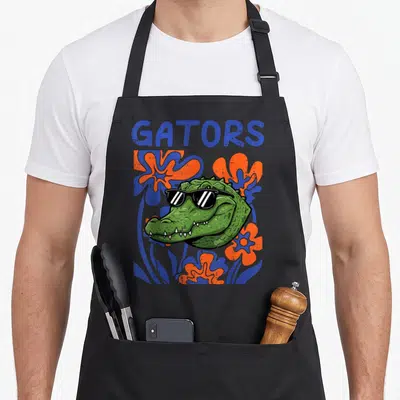 Youth floral apron - Cartoon alligator head with sunglasses surrounded by blue and orange flowers under GATORS text — shown on a black cotton canvas apron against a white background