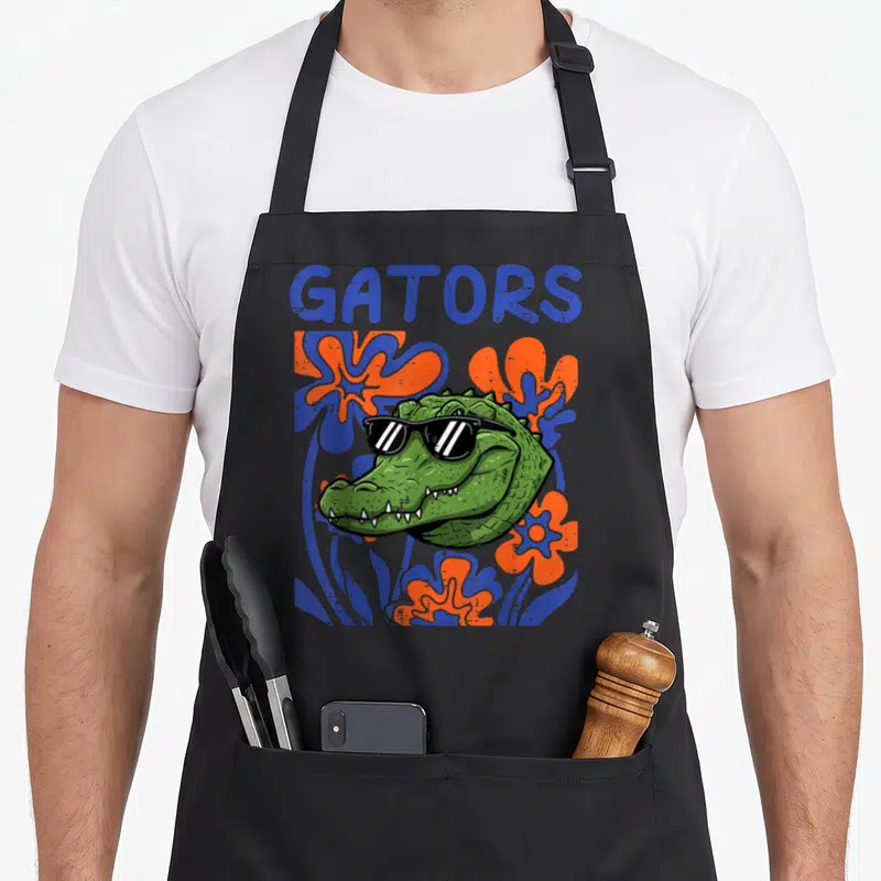 Youth floral apron - Cartoon alligator head with sunglasses surrounded by blue and orange flowers under GATORS text — shown on a black cotton canvas apron against a white background