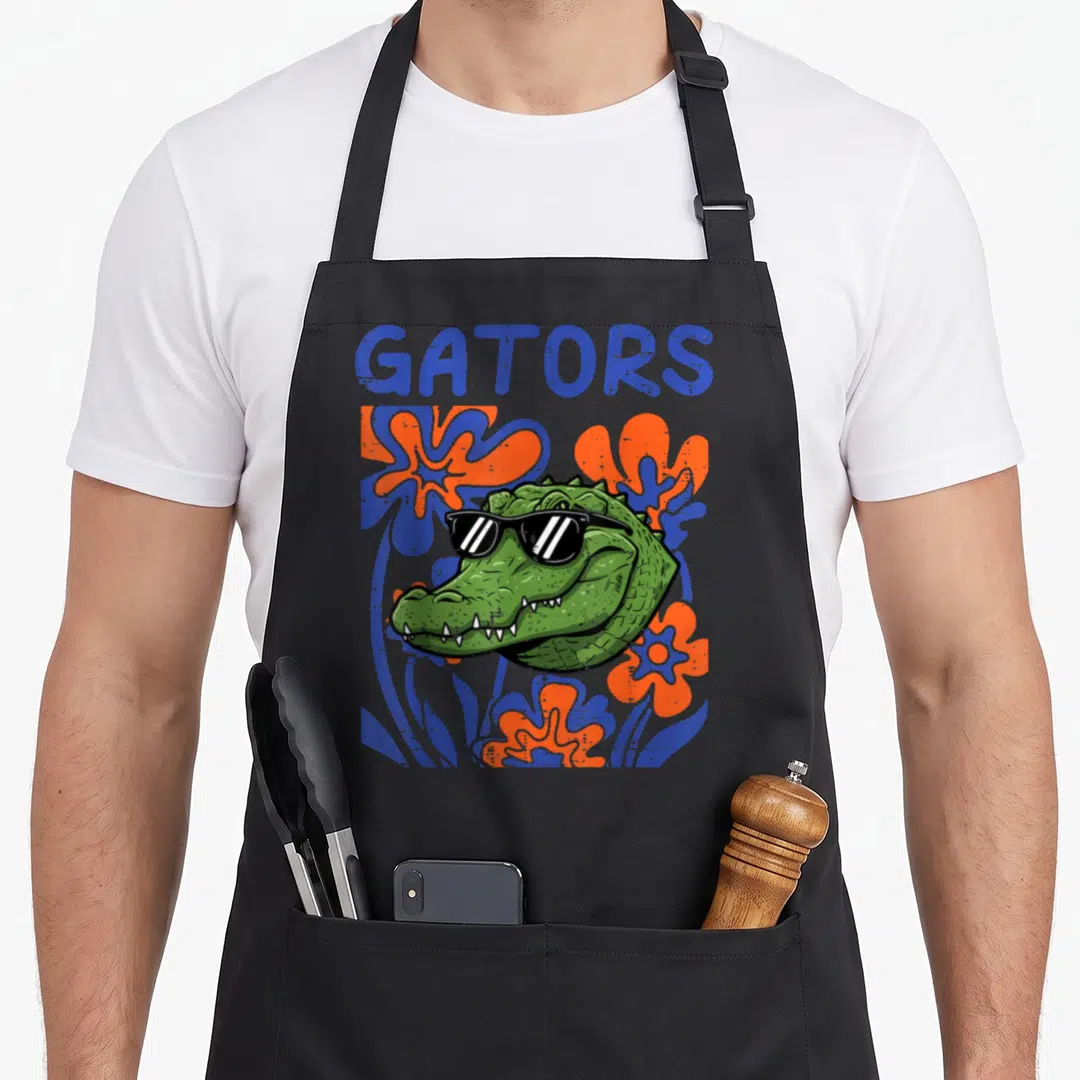Youth floral apron - Cartoon alligator head with sunglasses surrounded by blue and orange flowers under GATORS text — shown on a black cotton canvas apron against a white background