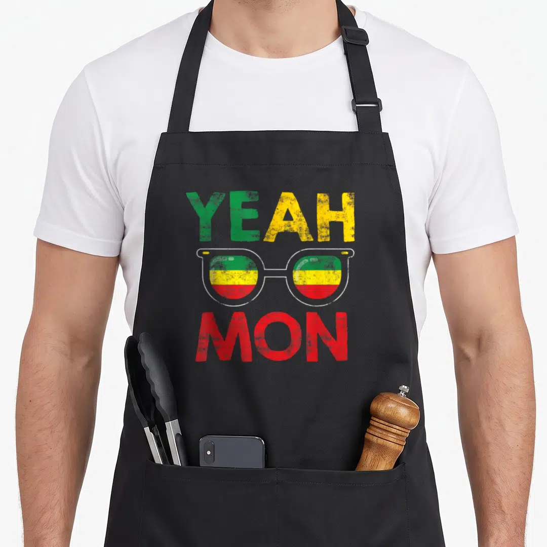 Mon yeah love apron - YEAH MON text with glasses containing green yellow red and white stripes — shown on a black cotton canvas apron against a white background