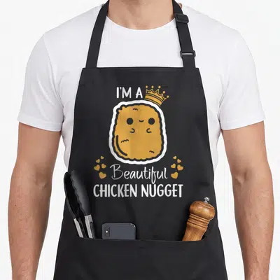 Beautiful chicken print apron - Cartoon golden nugget wearing a crown with text I'M A Beautiful CHICKEN NUGGET — shown on a black cotton canvas apron against a white background