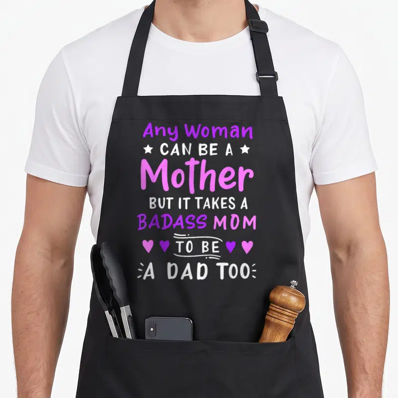 Cute single mother's day aprons - Purple and pink stacked text reading Any Woman Can Be A Mother But It Takes A Badass — shown on a black cotton canvas apron against a white background
