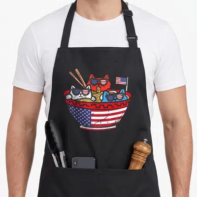 Ramen 4th of july apron - Three cats in sunglasses sitting in a ramen bowl with an American flag and chopsticks — shown on a black cotton canvas apron against a white background