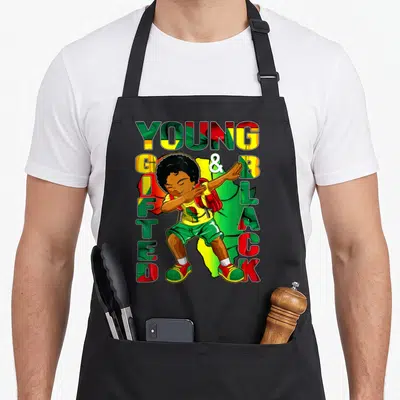 History gifted apron - Cartoon boy dabbing surrounded by YOUNG GIFTED & BLACK text in red green and yellow patterns — shown on a black cotton canvas apron against a white background