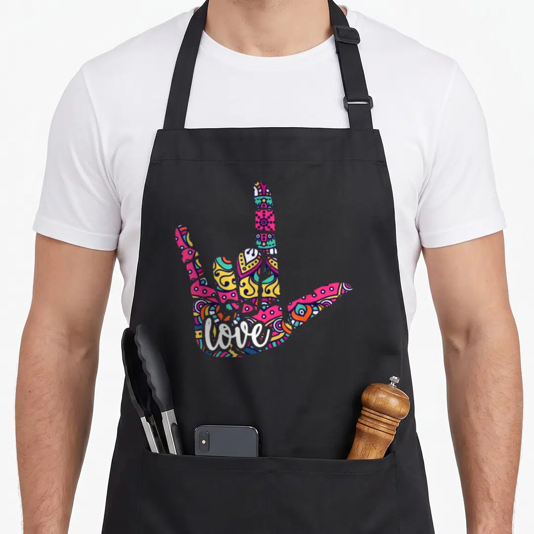Funny sign language apron - Multicolored hand making I love you sign with white love script on palm — shown on a black cotton canvas apron against a white background