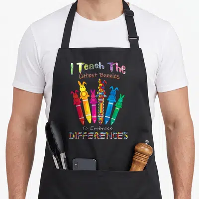 Autism teacher apron - Six bunny crayons in red orange pink blue green yellow with puzzle pieces and text I Teach — shown on a black cotton canvas apron against a white background