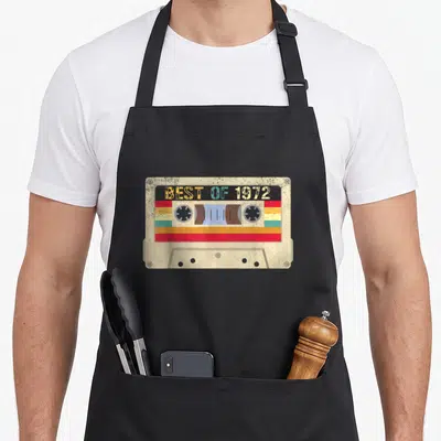 Decoration bday birthday apron - Retro cassette tape with BEST OF 1972 text and multicolor horizontal stripes — shown on a black cotton canvas apron against a white background