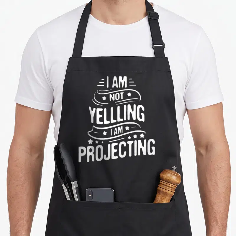 Projecting theatre apron - White text I AM NOT YELLLING I AM PROJECTING with ribbons and stars — shown on a black cotton canvas apron against a white background