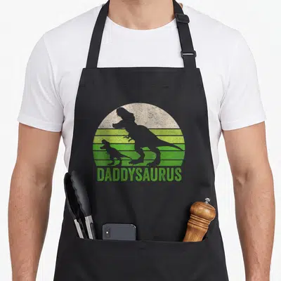 Daddy daddysaurus dinosaur apron - Grey T-Rex silhouettes against striped green and beige sun with DADDYSAURUS text — shown on a black cotton canvas apron against a white background