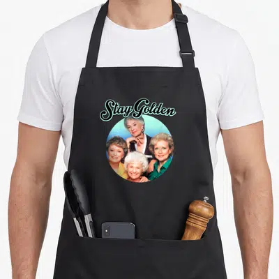 Science golden apron - Stay Golden text above circular photo of four smiling women in blue and green — shown on a black cotton canvas apron against a white background