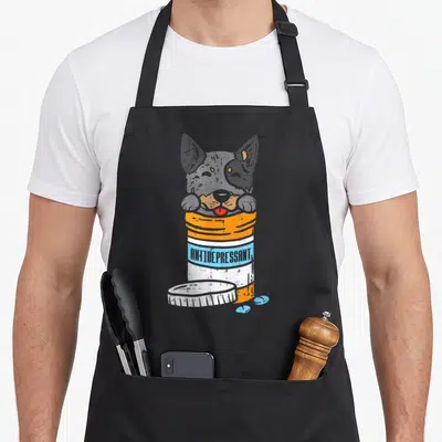 Antidrepressant dog apron - Heeler head inside pill bottle with ANTIDEPRESSANT text in orange and blue distressed print — shown on a black cotton canvas apron against a white background