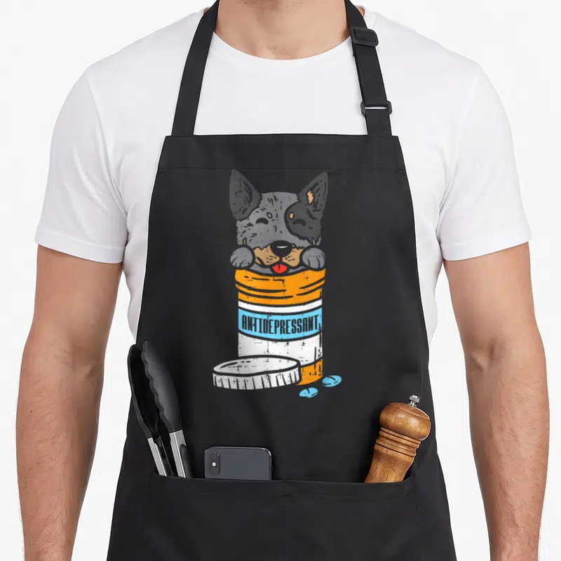 Antidrepressant dog apron - Heeler head inside pill bottle with ANTIDEPRESSANT text in orange and blue distressed print — shown on a black cotton canvas apron against a white background