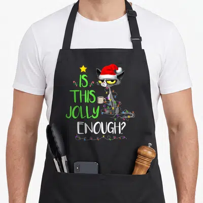 Jolly christmas tree apron - Grey cartoon cat in Santa hat wrapped in lights next to text IS THIS JOLLY ENOUGH — shown on a black cotton canvas apron against a white background