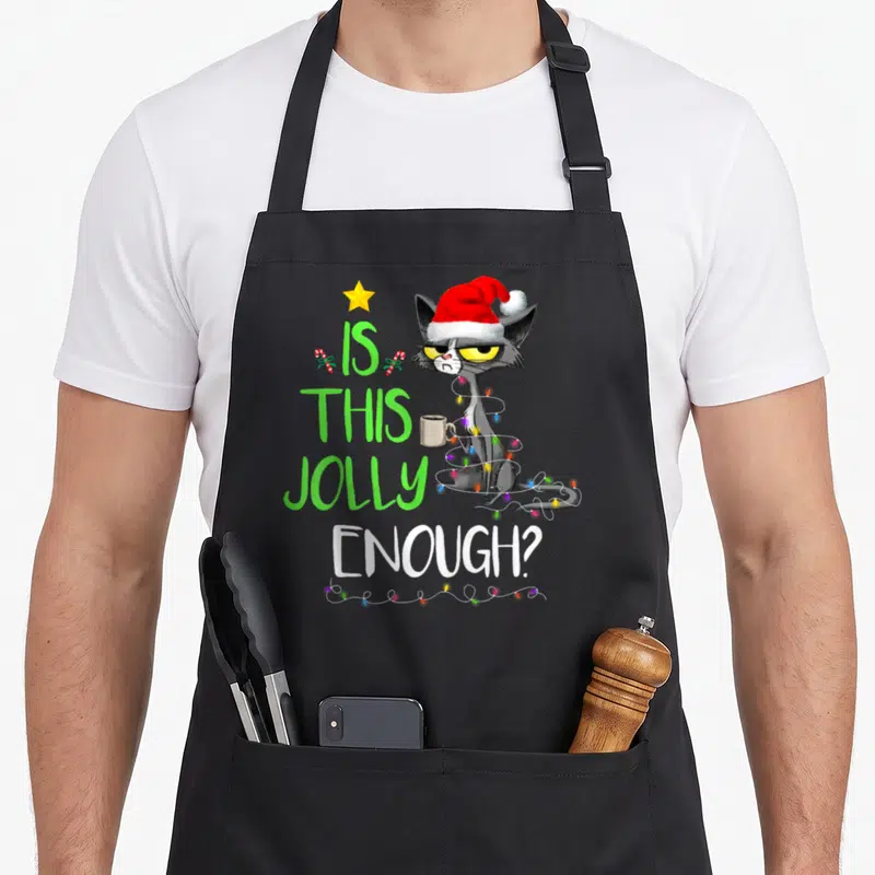 Jolly christmas tree apron - Grey cartoon cat in Santa hat wrapped in lights next to text IS THIS JOLLY ENOUGH — shown on a black cotton canvas apron against a white background