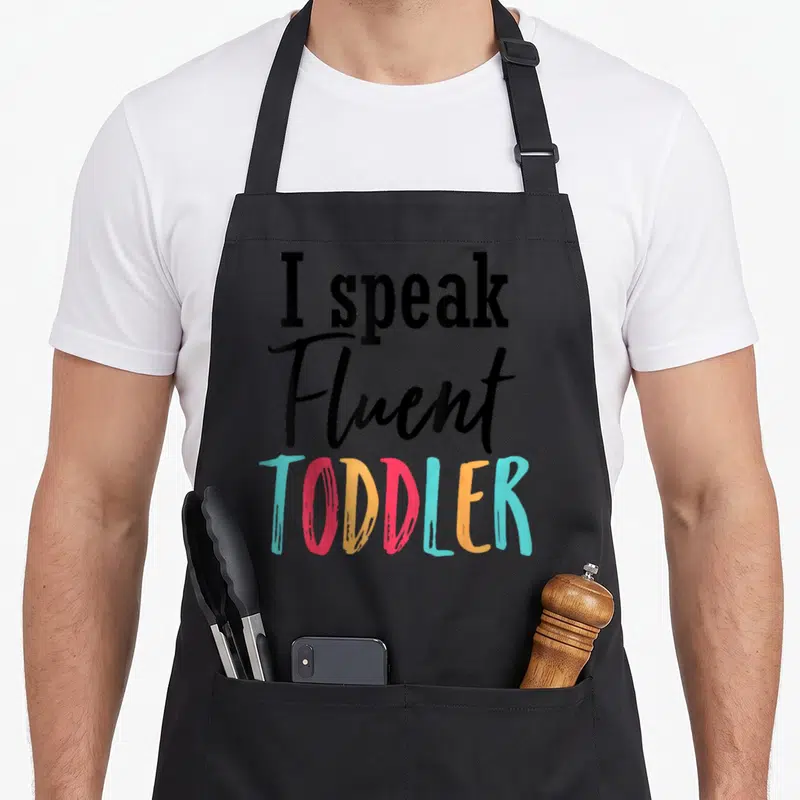 Speak preschool teacher apron - I speak Fluent TODDLER text in black script and multicolor block letters — shown on a black cotton canvas apron against a white background