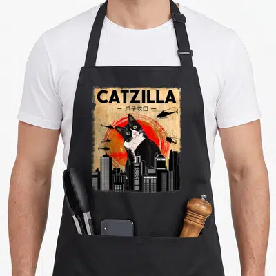 Catzilla tuxedo apron - Giant tuxedo cat in front of red sun with helicopters and black skyline — shown on a black cotton canvas apron against a white background