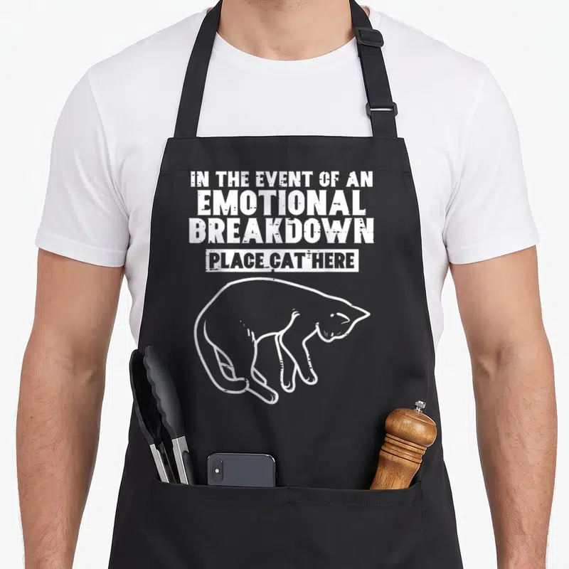 Event cat apron - White stacked text reading IN THE EVENT OF AN EMOTIONAL BREAKDOWN PLACE CAT HERE above a line drawing — shown on a black cotton canvas apron against a white background