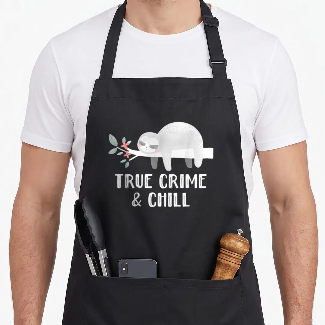 Crime sloth apron - Cartoon sloth lying on branch with flowers above TRUE CRIME & CHILL text — shown on a black cotton canvas apron against a white background