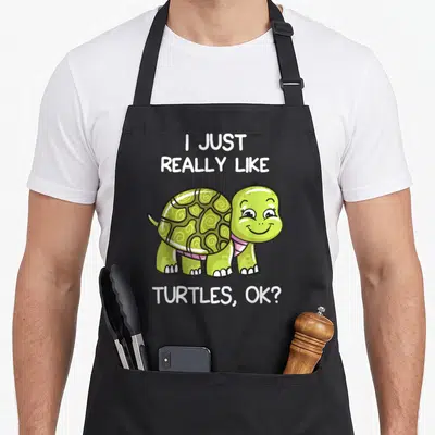 Cute turtle lover apron - Cartoon green turtle with pink collar and text I Just Really Like Turtles Ok — shown on a black cotton canvas apron against a white background