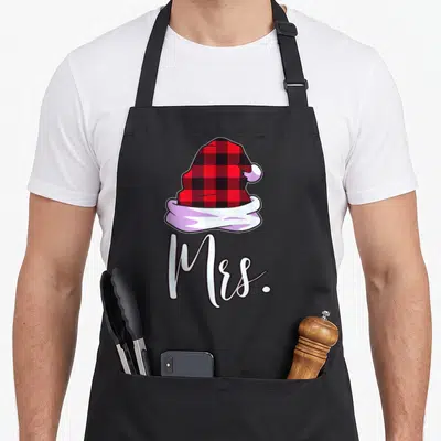 Plaid santa apron - Red and black plaid Santa hat illustration above white Mrs script text — shown on a black cotton canvas apron against a white background