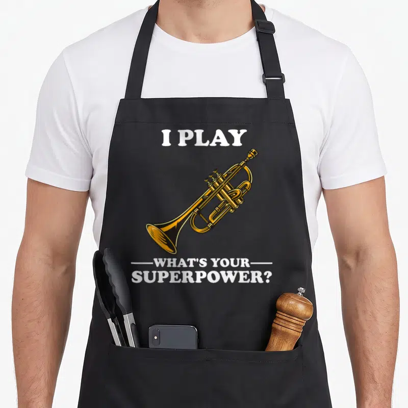Motif trumpet apron - Gold cartoon trumpet with white text I Play What's Your Superpower flanked by lines — shown on a black cotton canvas apron against a white background