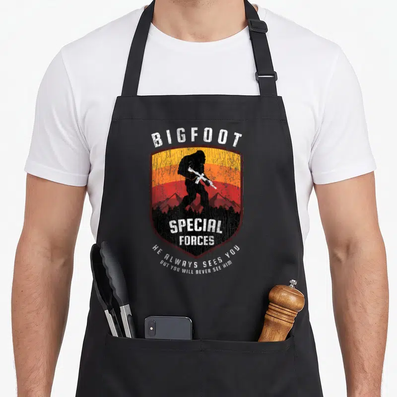 Bigfoot special aprons - Retro shield with Bigfoot silhouette, orange stripes, and text He Always Sees You But You Will — shown on a black cotton canvas apron against a white background
