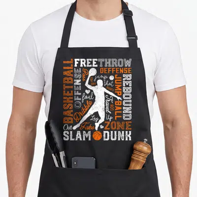 Basketball production life apron - White player silhouette surrounded by orange and white basketball terms like SLAM — shown on a black cotton canvas apron against a white background