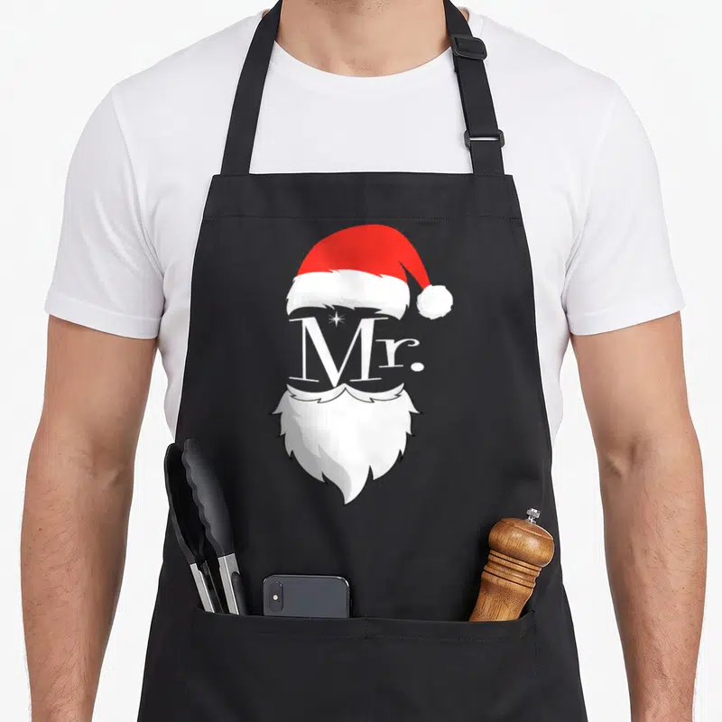 Claus mr mrs aprons - Red Santa hat, white Mr text, and white beard stacked vertically on black background — shown on a black cotton canvas apron against a white background
