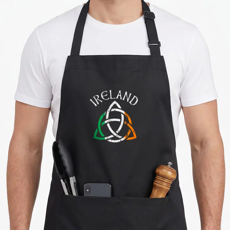 Ireland st patrick day aprons - Distressed triquetra knot in green white orange with arched IRELAND text above — shown on a black cotton canvas apron against a white background