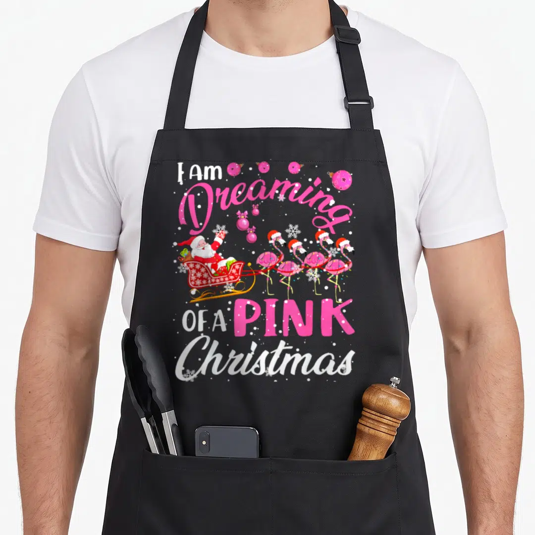 Dreaming pink flamingo apron - Santa sleigh pulled by four pink flamingos with pink and white holiday text — shown on a black cotton canvas apron against a white background