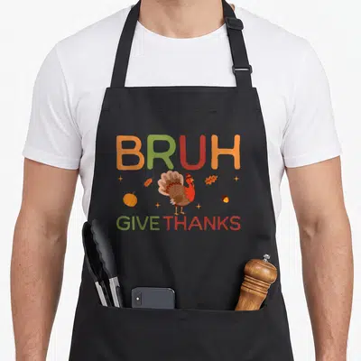 Bruh thankful apron - Cartoon turkey wearing a hat surrounded by pumpkins and leaves under BRUH GIVE THANKS text — shown on a black cotton canvas apron against a white background
