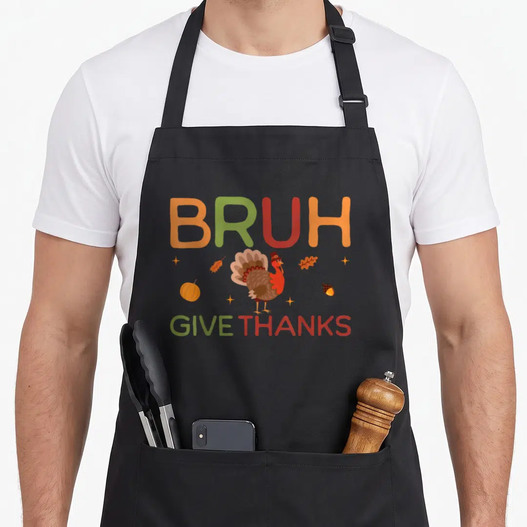 Bruh thankful apron - Cartoon turkey wearing a hat surrounded by pumpkins and leaves under BRUH GIVE THANKS text — shown on a black cotton canvas apron against a white background