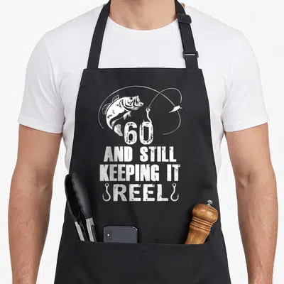 Keeping 60th birthday apron - White fish jumping above 60 text and fishing hooks on black apron — shown on a black cotton canvas apron against a white background