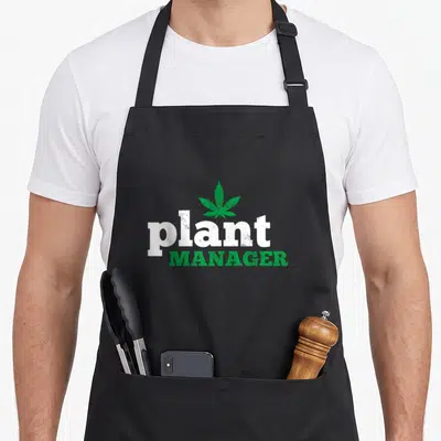 Thc plant apron - Green cannabis leaf above white plant and green MANAGER text in distressed style — shown on a black cotton canvas apron against a white background