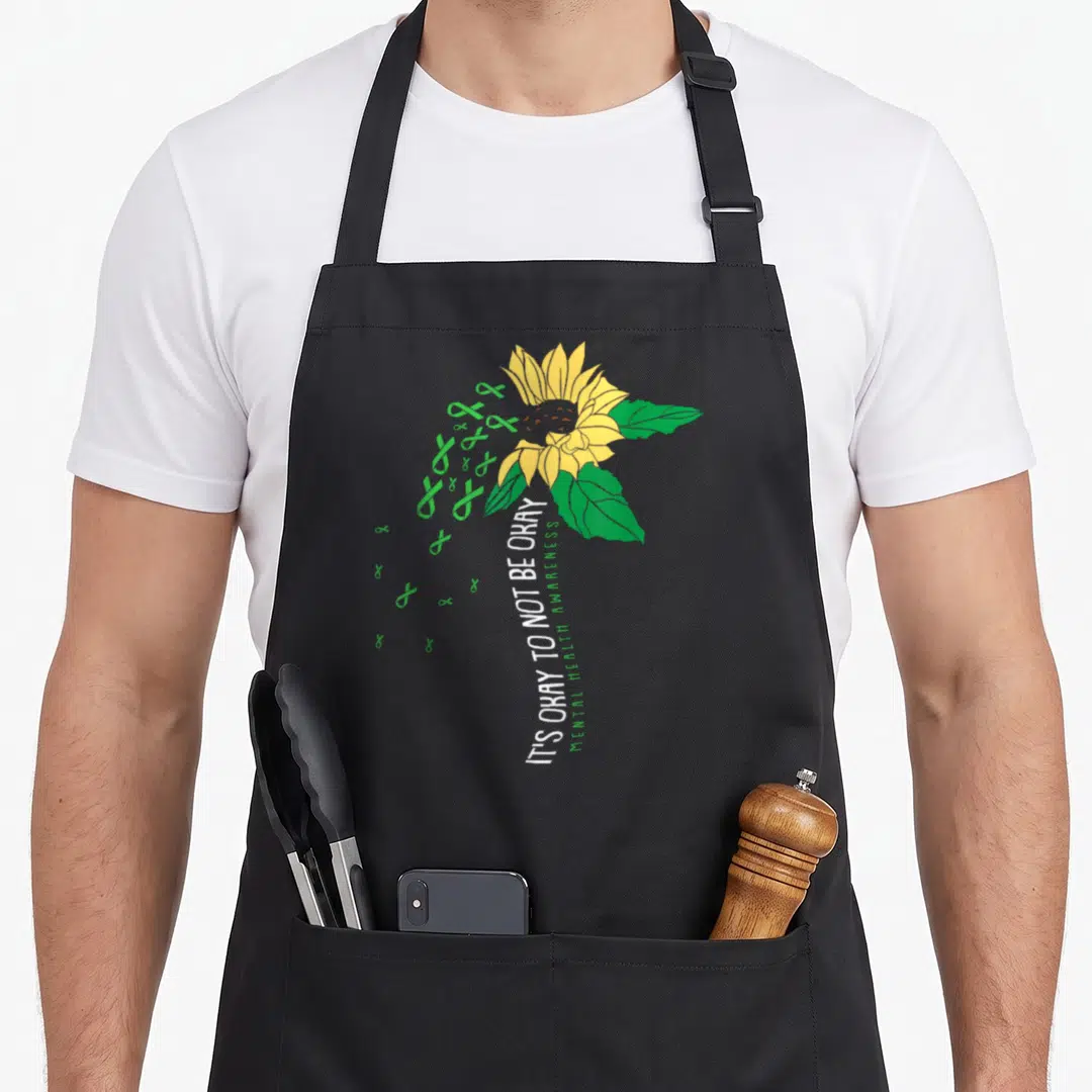 Awareness sunflower apron - Yellow sunflower with green ribbons and vertical white text IT'S OKAY TO NOT BE OKAY — shown on a black cotton canvas apron against a white background