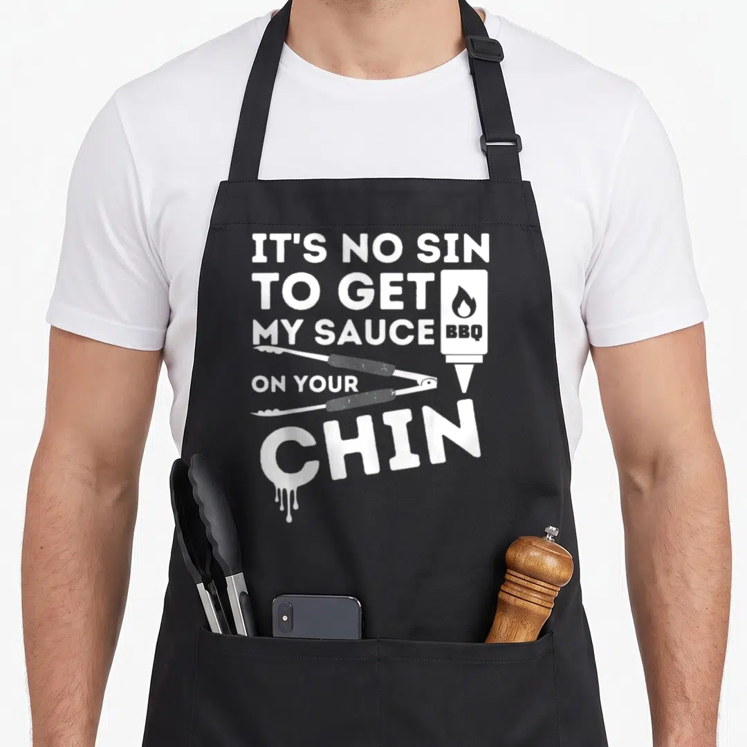 Sin barbecue apron - White text IT'S NO SIN TO GET MY SAUCE ON YOUR CHIN with sauce bottle, tongs, and dripping C — shown on a black cotton canvas apron against a white background