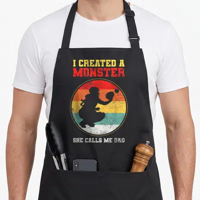 Catcher father day apron - I CREATED A MONSTER SHE CALLS ME DAD text with softball catcher silhouette on striped circle — shown on a black cotton canvas apron against a white background