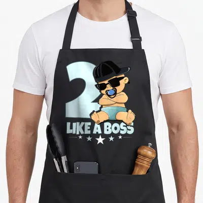 Baby 2nd bbq boss apron - Cartoon baby with sunglasses and hat before light blue 2, LIKE A BOSS text and stars — shown on a black cotton canvas apron against a white background