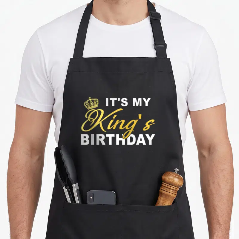 Birthday couple kings apron - Gold crown next to stacked IT'S MY King's BIRTHDAY text in white and gold typography — shown on a black cotton canvas apron against a white background
