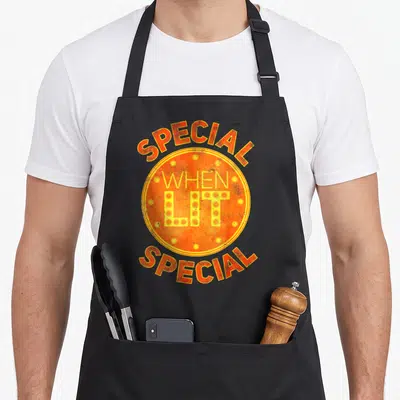 Lit special aprons - Retro orange circle with SPECIAL WHEN LIT text and yellow dots — shown on a black cotton canvas apron against a white background