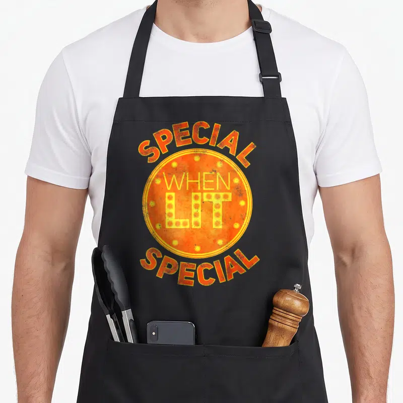 Lit special aprons - Retro orange circle with SPECIAL WHEN LIT text and yellow dots — shown on a black cotton canvas apron against a white background