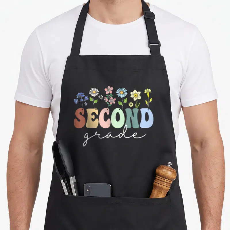 2nd floral apron - SECOND grade text with cartoon wildflowers in orange yellow pink green blue — shown on a black cotton canvas apron against a white background
