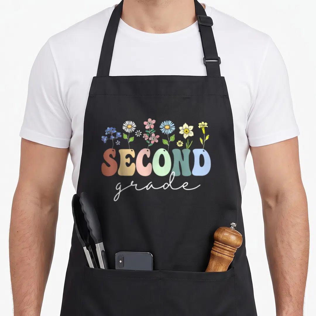 2nd floral apron - SECOND grade text with cartoon wildflowers in orange yellow pink green blue — shown on a black cotton canvas apron against a white background
