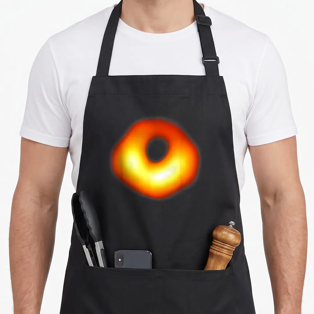 Funny hole apron - Glowing orange and yellow ring with dark center representing a black hole — shown on a black cotton canvas apron against a white background