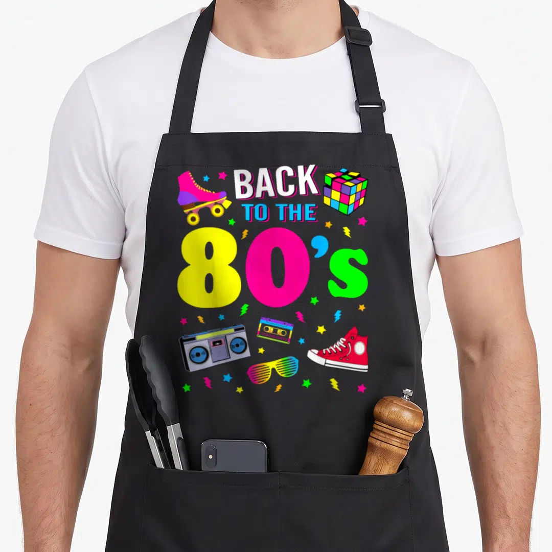 Eighty 80s apron - Back to the 80s text with cartoon boombox, roller skate, Rubik's cube, and colorful stars scattered — shown on a black cotton canvas apron against a white background