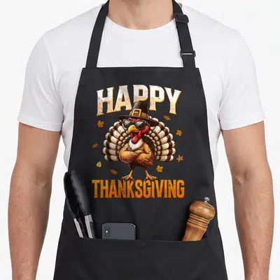 Retro family thanksgiving apron - Cartoon turkey in sunglasses and hat between HAPPY THANKSGIVING text with scattered — shown on a black cotton canvas apron against a white background