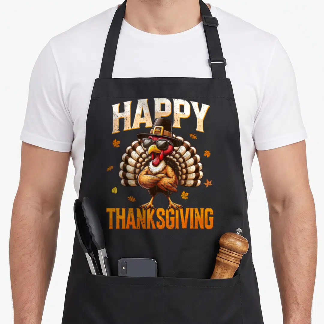 Retro family thanksgiving apron - Cartoon turkey in sunglasses and hat between HAPPY THANKSGIVING text with scattered — shown on a black cotton canvas apron against a white background