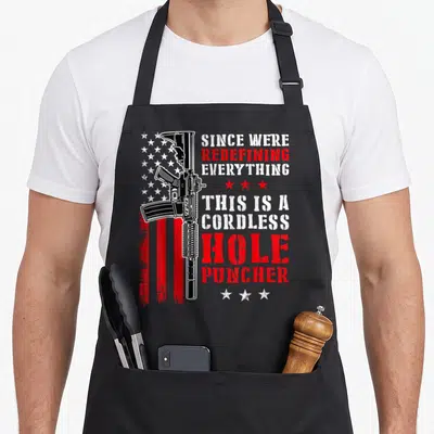 Redefine hole apron - Vertical rifle over distressed flag with red and white text reading redefine hole puncher — shown on a black cotton canvas apron against a white background