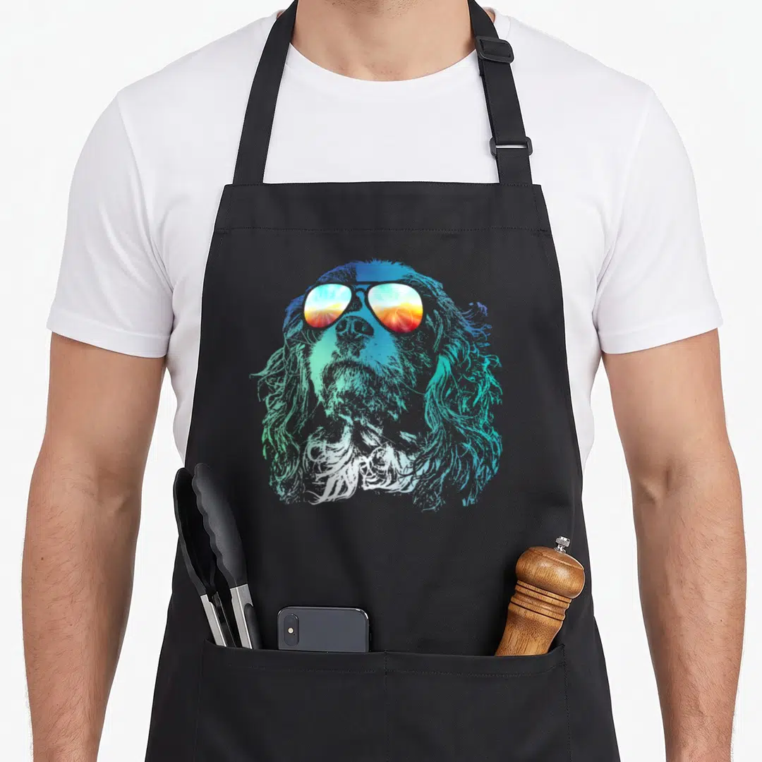 Groovy king apron - Realistic dog head in blue and cyan gradients wearing sunglasses reflecting an orange and red sunset — shown on a black cotton canvas apron against a white background